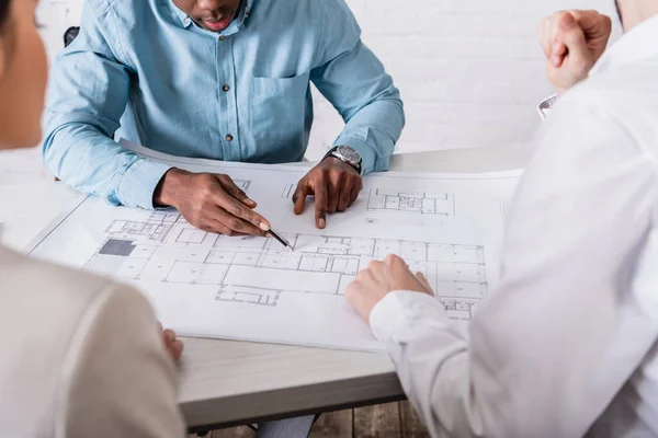 Partial view of african american businessman pointing with pencil at blueprint near multiethnic business partners,  blurred foreground — Stock Photo