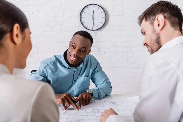 Smiling african american businessman pointing at blueprint while looking at business partner near translator on blurred foreground — Stock Photo