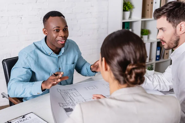 Animado empresário afro-americano gesticular enquanto conversa com parceiro de negócios e intérprete perto do projeto, foreground borrado — Fotografia de Stock