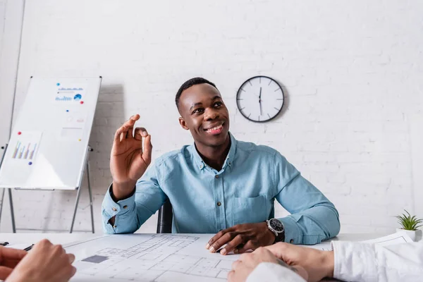 Cheerful african american businessman showing okay gesture near blueprint and business partners on foreground — Stock Photo