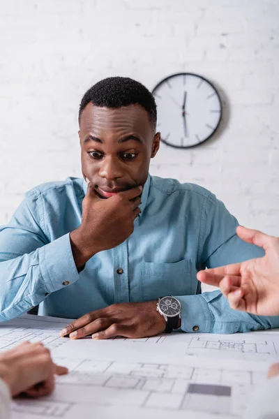 Desanimado hombre de negocios afroamericano mirando el proyecto cerca de socios de negocios que hacen gestos en primer plano borroso - foto de stock