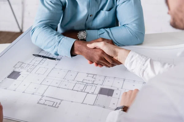 Cropped view of african american businessman shaking hands with business partner near blueprint, blurred foreground — Stock Photo