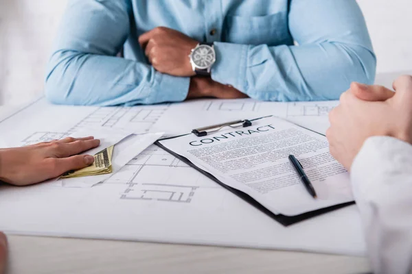 Cropped view of woman giving bribe to african american businessman near blueprint and contract, blurred background — Stock Photo