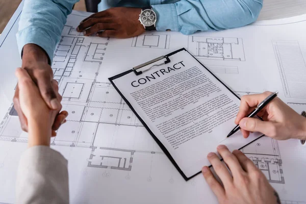 Cropped view of businessman signing contract near interracial partners shaking hands near blueprint — Stock Photo