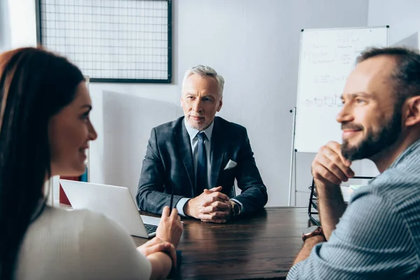 Mature investor looking at smiling businesspeople talking on blurred foreground in office — Stock Photo