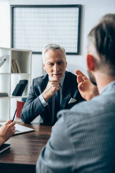 Investor in formal wear looking at businessman on blurred foreground near laptop in office — Stock Photo