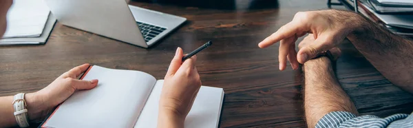 Vista ritagliata dell'uomo d'affari che punta con il dito vicino al collega con penna, notebook e laptop su sfondo sfocato sul tavolo dell'ufficio, banner — Foto stock