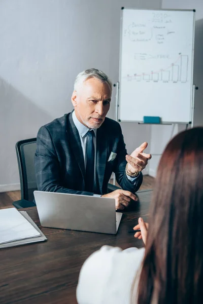 Reifer Investor zeigt mit der Hand in die Nähe von Laptop, Papieren und Geschäftsfrau auf verschwommenem Vordergrund — Stockfoto