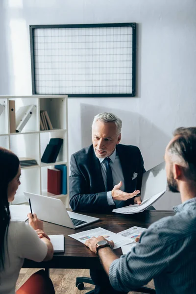 Inversor sonriente señalando con la mano en la carpeta de papel cerca de los empresarios en primer plano borroso y portátil en la mesa — Stock Photo