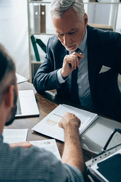 Pensive investor looking at papers with graphs near laptop and businessman on blurred foreground — Stock Photo