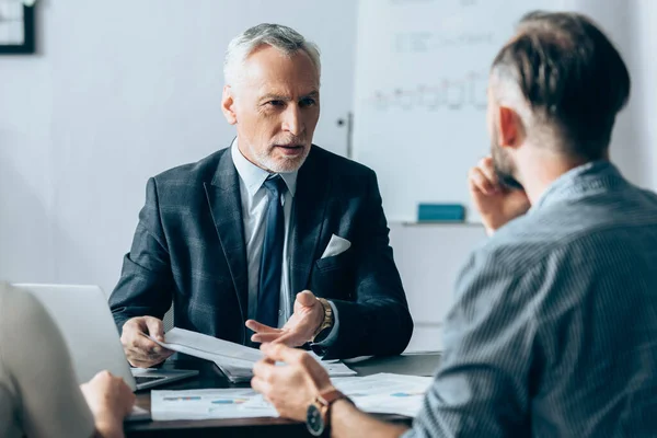 Investisseur pointant vers les papiers près des gens d'affaires et ordinateur portable sur le premier plan flou — Photo de stock