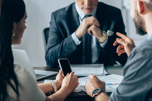 Empresario señalando con el dedo cerca colega con teléfono inteligente e inversor en fondo borroso en la oficina — Stock Photo