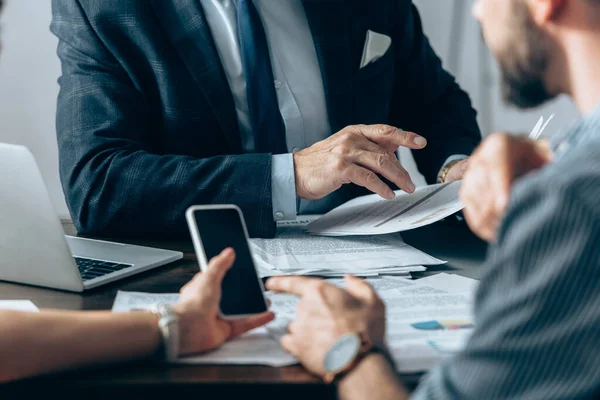 Cropped view of investor holding document near business people pointing at smartphone on blurred foreground in office — Stock Photo