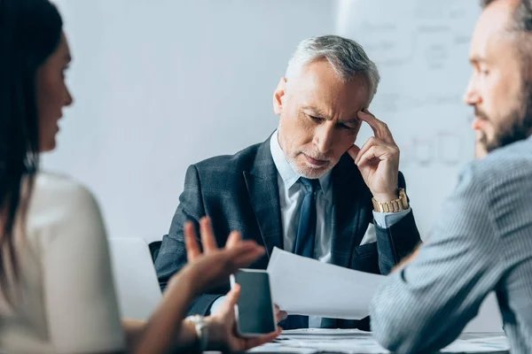 Mature investor looking at documents while business people with smartphone talking on blurred foreground — Stock Photo