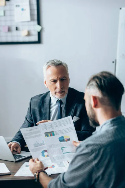 Investisseur utilisant un ordinateur portable et regardant un homme d'affaires avec des documents au premier plan flou dans le bureau — Photo de stock