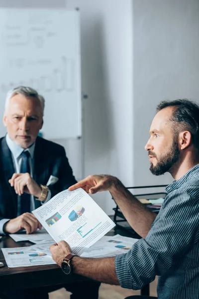 Бизнесмен держит бумагу с графиками рядом с инвестором на размытом фоне в офисе — стоковое фото
