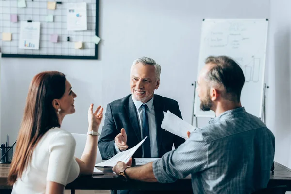 Lächelnder Investor zeigt auf Dokumente in der Nähe von Geschäftsleuten im verschwommenen Vordergrundgespräch im Büro — Stockfoto