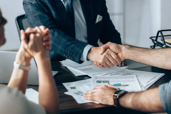 Vista recortada del hombre de negocios estrechando la mano con el inversor cerca de colega y papeles en la mesa - foto de stock