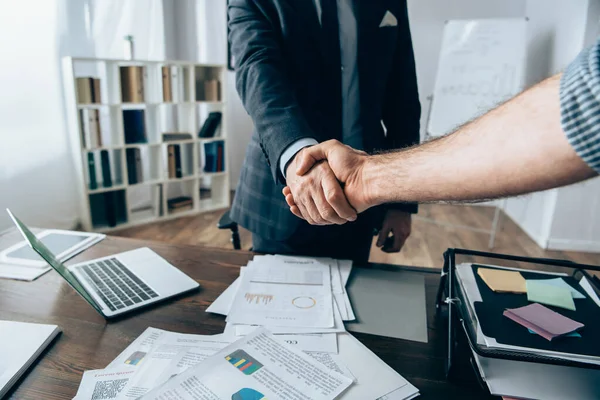 Vista recortada del empresario estrechando la mano del inversor cerca del papeleo con gráficos y dispositivos sobre la mesa - foto de stock