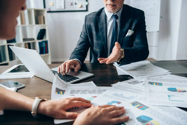 Vista recortada del inversor utilizando el ordenador portátil y apuntando con la mano cerca de la mujer de negocios con papeles en primer plano borrosa - foto de stock