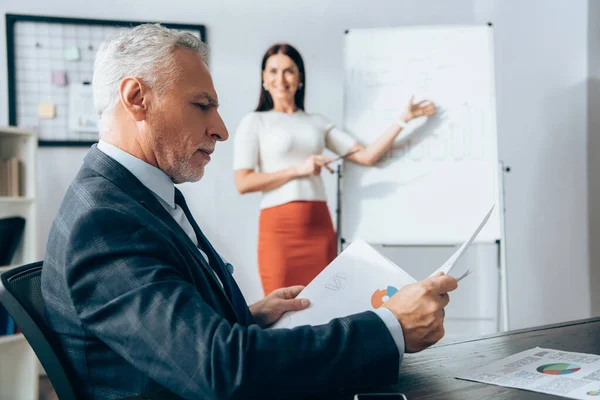 Reifer Investor betrachtet Dokumente in der Nähe einer Geschäftsfrau, die neben Flipchart auf verschwommenem Hintergrund im Büro steht — Stockfoto