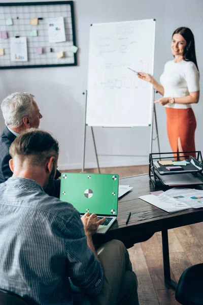 Uomo d'affari che utilizza laptop con schermo verde vicino investitore e collega che punta a flipchart su sfondo sfocato — Foto stock