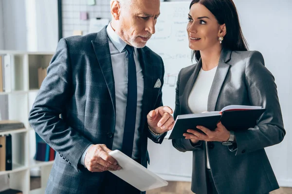 Smiling businesswoman holding notebook near investor with digital tablet pointing with finger — Stock Photo