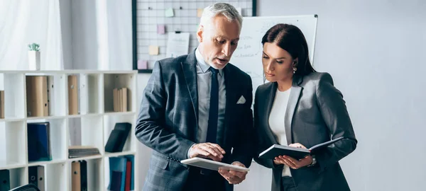 Mature investor using digital tablet near businesswoman with notebook in office, banner — Stock Photo