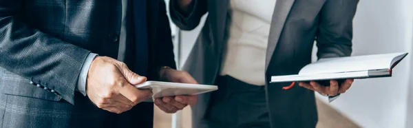 Обрізаний вид консультанта, який тримає цифровий планшет біля бізнес-леді з блокнотом на розмитому фоні в офісі, банер — стокове фото