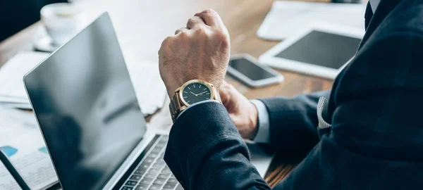 Ausgeschnittene Ansicht der Armbanduhr auf der Hand des Geschäftsmannes, der neben Laptop mit leerem Bildschirm sitzt, Banner — Stockfoto