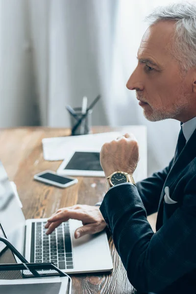 Investitore in tuta utilizzando laptop vicino a dispositivi su sfondo sfocato in ufficio — Foto stock