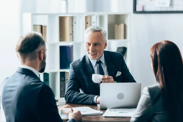 Investisseur souriant avec une tasse de café regardant les gens d'affaires sur le premier plan flou près de papiers et ordinateur portable — Photo de stock