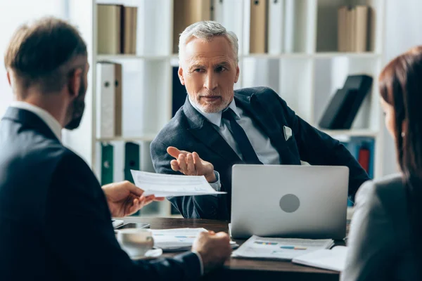 Reifer Investor zeigt mit Dokument auf verschwommenen Vordergrund neben Papieren und Laptop auf Geschäftsmann — Stockfoto