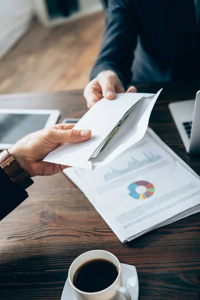 Ausgeschnittene Ansicht eines Investors auf verschwommenem Hintergrund, der Geschäftsmann in der Nähe von Papieren und einer Tasse Kaffee einen Umschlag mit Geld gibt — Stockfoto