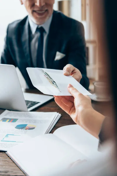 Cropped view of businesswoman taking envelope with money from investor near papers and laptop on blurred background — Stock Photo