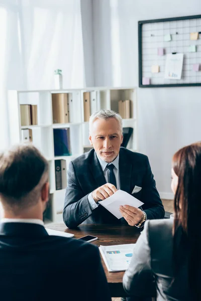 Mature advisor holding envelope near business people on blurred foreground — Stock Photo