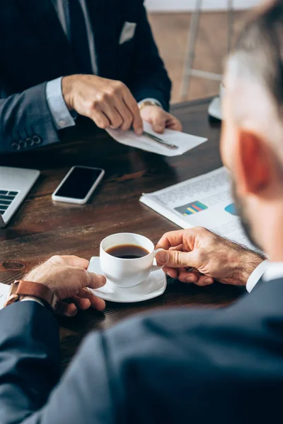 Coupe de café dans les mains de l'homme d'affaires près de l'investisseur avec enveloppe et papiers sur fond flou — Photo de stock