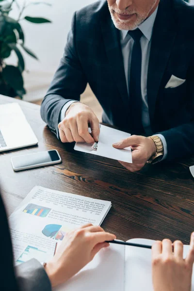 Cropped view of businessman holding envelope and money near businesswoman with pen and papers with graphs — Stock Photo