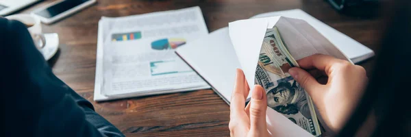 Ausgeschnittene Ansicht einer Geschäftsfrau mit Dollars und Umschlag in der Nähe von Papieren auf verschwommenem Hintergrund im Büro, Banner — Stockfoto