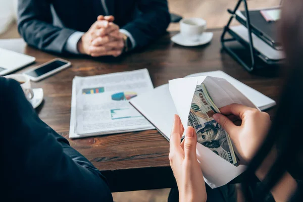 Vista ritagliata di donna d'affari in possesso di dollari e busta vicino investitore e documenti su sfondo sfocato — Foto stock