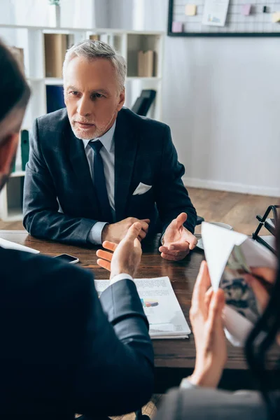 Reifer Investor im Gespräch mit Geschäftsmann nahe Kollege mit Geld und Umschlag im verschwommenen Vordergrund — Stockfoto