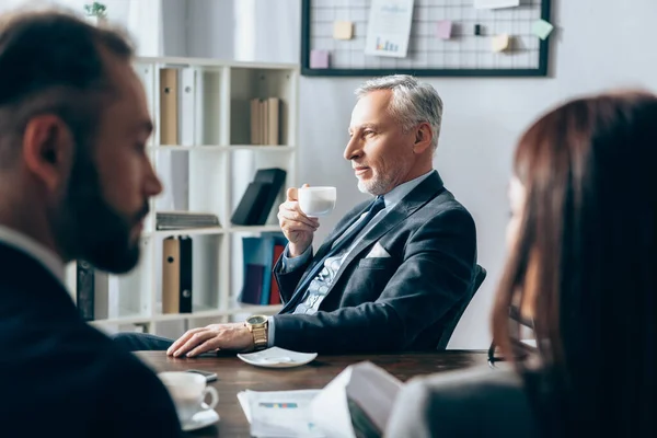 Mature investor holding coffee cup near business people on blurred foreground in office — Stock Photo