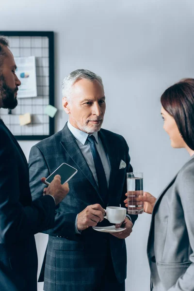 Businesswoman with glass of water standing near businessmen with coffee and smartphone in office — Stock Photo