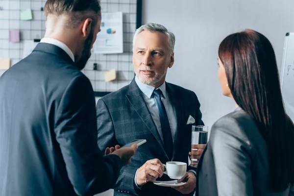 Mature advisor with cup  of coffee looking at businessman with smartphone and businesswoman with glass of water on blurred foreground — Stock Photo