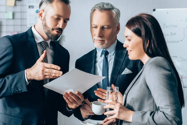 Homme d'affaires pointant vers le papier près souriant femme d'affaires avec de l'eau et investisseur avec du café dans le bureau — Photo de stock