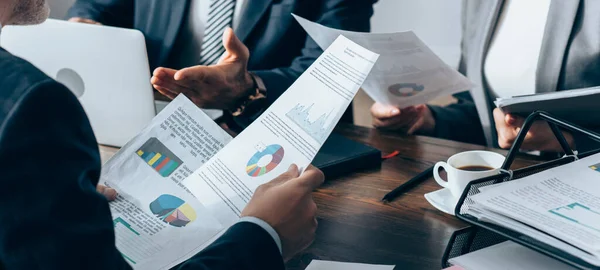 Ausgeschnittene Ansicht eines Beraters, der Papiere mit Diagrammen in der Nähe von Geschäftsleuten hält, Laptop und Notizbuch auf dem Tisch, Banner — Stockfoto