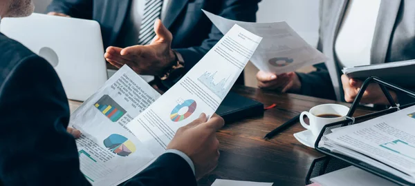 Cropped view of advisor holding papers with charts near business people, laptop and notebook on table, banner — Stock Photo