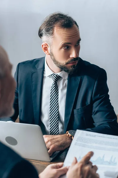 Businessman using laptop near advisor pointing at paper on blurred foreground — Stock Photo