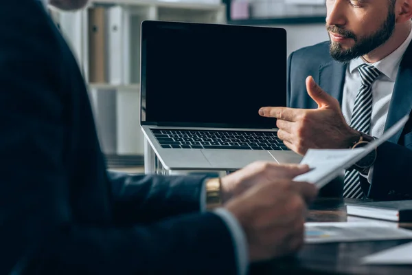 Vista ritagliata dell'uomo d'affari che punta al laptop con schermo vuoto vicino al consulente con carta in primo piano sfocata — Foto stock