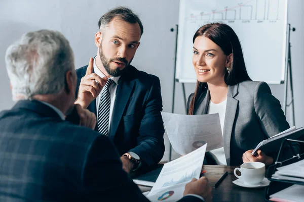 Geschäftsmann zeigt mit Finger auf lächelnde Geschäftsfrau mit Dokumenten und Investor im verschwommenen Vordergrund — Stockfoto